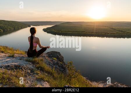 Woman in pink top and black leggings is practicing yoga performing yoga-lotus position outdoors, near a riverside Stock Photo