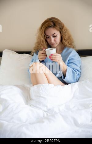 Woman in blue sweater with the cup of tea sitting in the bed. Stock Photo
