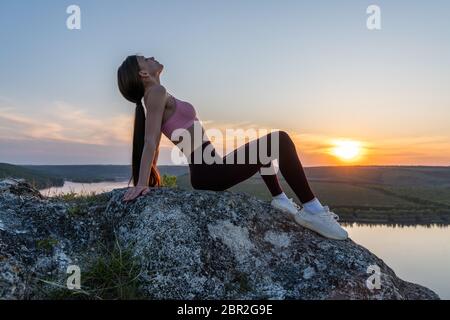 Young healthy woman in stylish sportswear practice yoga on mountain peak cliff Stock Photo