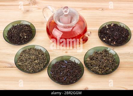 Various kinds of dry tea in glass saucers and teapot with hot tea standing on a wooden background.  Different kinds of tea leaves. Stock Photo