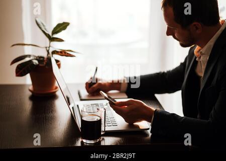 Businessman making notes and using gadgets phone and laptop. Office worker writing in notebook and using laptop with plant and window background. Mana Stock Photo