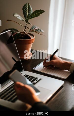Businessman making notes and using gadgets. Office worker writing in notebook and using laptop with plant and window background. Manager Handwriting d Stock Photo