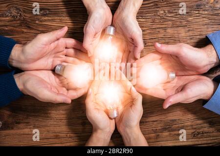 An Elevated View Of Businesspeople's Hand Holding Illuminated Light Bulbs In Hands Over Wooden Desk Stock Photo