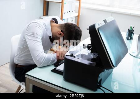 Tired Businessman Resting His Head In Front Of Paper Stuck In Printer Stock Photo