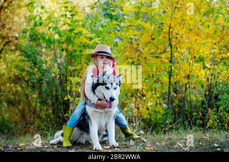 Girl in hat hugging her dog. Little girl with her dog, alaskan malamute, full length. Child and dog on nature background. Carefree childhood. Nature w Stock Photo