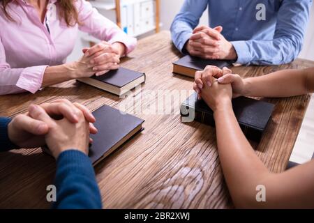 Close-up Of Businesspeople's Praying Hands Over The Bible On Wooden Desk Stock Photo