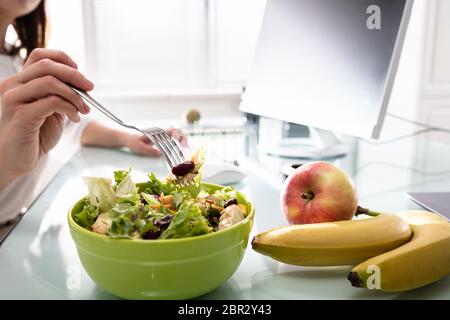 Close-up Of Woman's Hand Holding Fork While Having Healthy Salad On Office Desk Stock Photo