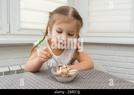 a beautiful 4-year-old girl in a white blouse eats ice-cream with chocolate from a snake bowl at a table in the kitchen with pleasure. baby is happy a Stock Photo