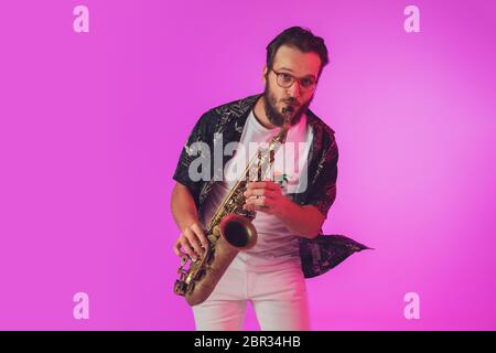 Young caucasian jazz musician playing the saxophone on gradient pink studio background in neon light. Concept of music, hobby, festival. Joyful, cheerful attractive guy. Colorful portrait of artist. Stock Photo