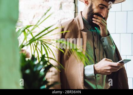 Image of joyful stylish young man wearing hat smiling and using cellphone in cafe Stock Photo