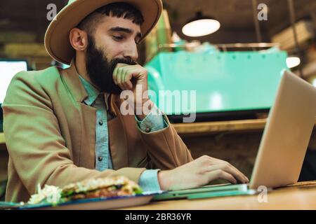 Image of attractive stylish young man wearing hat using laptop computer while having dinner in cafe Stock Photo