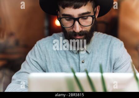 Photo of focused young man wearing eyeglasses using laptop while sitting at table in cafe indoors Stock Photo
