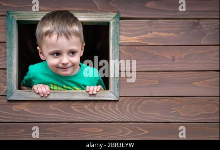 Portrait of a cute little Caucasian boy looking through the window of a wooden toy house in a outdoor playground Stock Photo