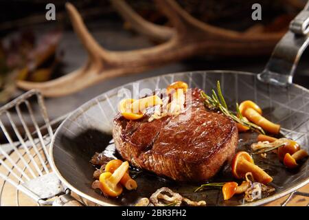 Gourmet thick marinated grilled wild venison steak with autumn mushrooms in a metal pan with blurred shed deer antler behind Stock Photo