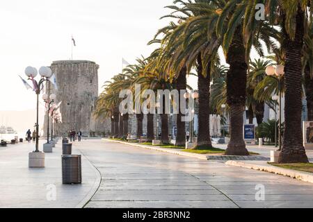 A few people stroll the palm tree lined Riva in the historic town of Trogir, Croatia. Kamerlengo Castle in the background. Stock Photo