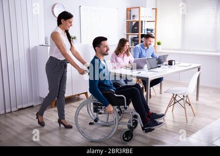 Smiling Young Businesswoman Helping Disabled Colleague In Wheelchair Stock Photo