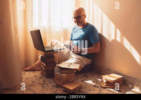 Middle-aged man working online with his laptop at home, sitting on the floor using a stack of books as a desk and using his smartphone. Concept work f Stock Photo