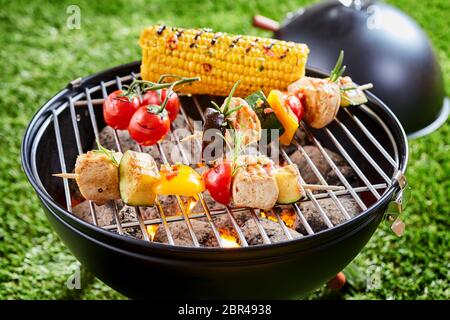 Vegetables and tofu kebab grilling on grid grille with fresh corn, viewed in close-up against green lawn grass in background Stock Photo