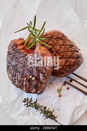 Two gourmet medallions of thick juicy grilled fillet beef steak seasoned with peppercorns and fresh thyme and rosemary on crumpled white paper, suitab Stock Photo