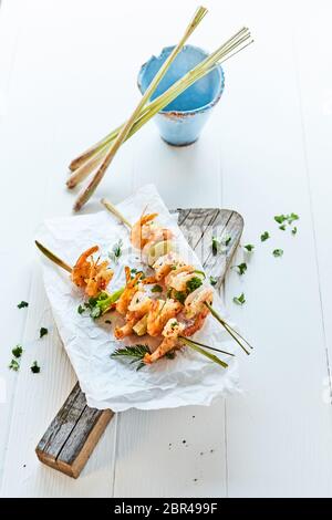 Gourmet grilled prawn tails seasoned with chilli spice and fresh herbs on bamboo skewers served on an old rustic wooden board on a white outdoor table Stock Photo