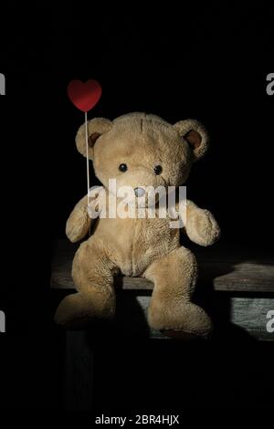 lonely brown teddy bear sits on a wooden chair and holds in its paw a red heart on a stick Stock Photo