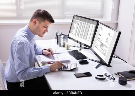 Young Businessman Calculating Invoice On Computer Screen Using Calculator Stock Photo