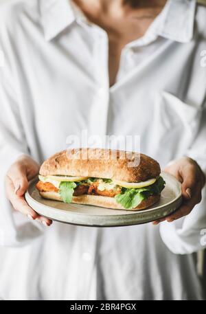 Woman in white shirt holding plate with fresh fried fish sandwich with tartare sauce, lemon and arugula, selective focus. Healthy easy breakfast ideas Stock Photo