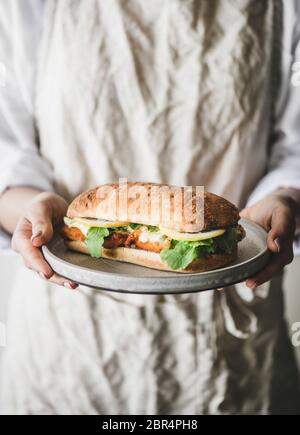 Woman in linen apron holding plate with fresh fried fish sandwich with tartare sauce, lemon and arugula, selective focus. Healthy easy breakfast ideas Stock Photo