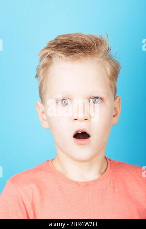 Portrait of cute surprised boy c with stylish hairstyle in coral T-shirt, closeup, on blue background. Stock Photo