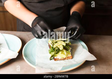 Cook hands preparing and making sandwich. Preparing healthy vegetarian bruschettas in dark gloves. Sandwich with soft cheese, arugula and cucumber Stock Photo