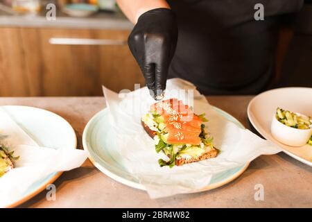 Cook hands preparing and making sandwich. Preparing healthy vegetarian bruschettas in dark gloves. Sandwich with soft cheese, arugula, cucumber and sa Stock Photo