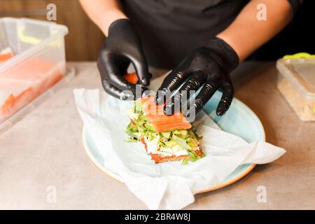 Cook hands preparing and making sandwich. Preparing healthy vegetarian bruschettas in dark gloves. Sandwich with soft cheese, arugula, cucumber and sa Stock Photo