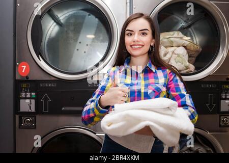 Girl pleased with the result of washing. Woman in laundromat showing thumbs up Stock Photo