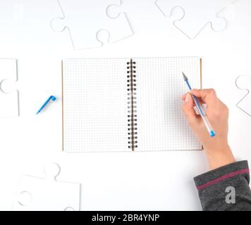 open notebook in a cell, hand holding a blue pen, next to it there are big white puzzles, top view Stock Photo