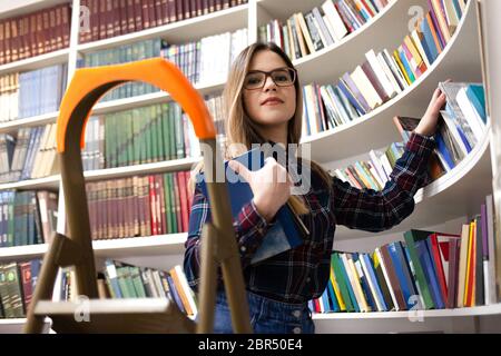 Cute student in glasses takes a book from the library bookshelf. Young lady is looking for the right literature among many books Stock Photo