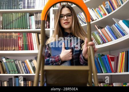 Pretty student lady stands on a stepladder in a library holding a book in her hand. Woman in public library Stock Photo