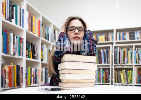 Young tired student girl sitting in a library at a table leaning on a large pile of books. Depressed woman in a plaid shirt prepares for exams in the Stock Photo