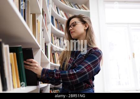 Cute student in glasses takes a book from the library bookshelf. Young lady is looking for the right literature among many books Stock Photo