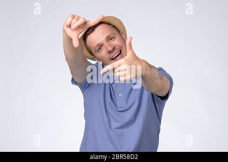 Curious man in summer hat gesturing picture frame with hands, looking through fingers and focusing on interesting moment, taking photo. Studio shot Stock Photo