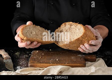 chef in a black uniform holds broken in half round rye bread over the table Stock Photo