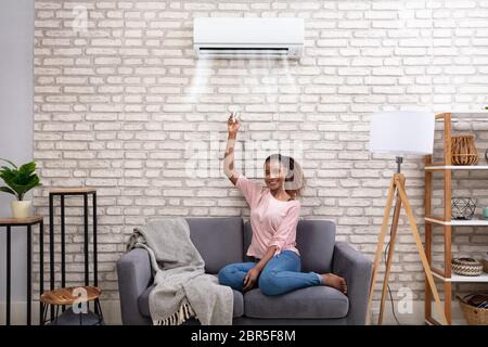 Young African Woman Switching On Air Conditioner With Remote Control At Home Stock Photo