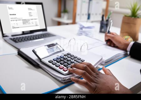 Close-up Of A Businesswoman Calculating Invoice Using Calculator Near Laptop Stock Photo