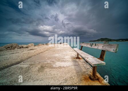 Empty wooden bench on a concrete pier with stormy dramatic clouds accumulating over Alykes Bay in Zante Island, Greece Stock Photo