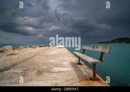 Empty wooden bench on a concrete pier with stormy dramatic clouds accumulating over Alykes Bay in Zante Island, Greece Stock Photo