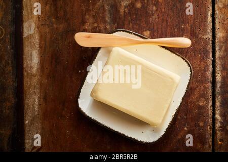 Whole pat of farm fresh creamy butter on a small rectangular butter dish with wooden spreader or knife viewed top down on a rustic wood table Stock Photo