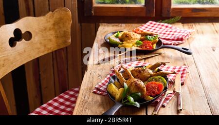 Serving of two crispy golden roast quails with roasted vegetables in a metal skillet at double place settings at a rustic table Stock Photo