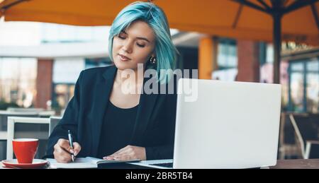 Cheerful lady with blue hair signing some documents in a restaurant using a laptop and drinking a cup of coffee Stock Photo