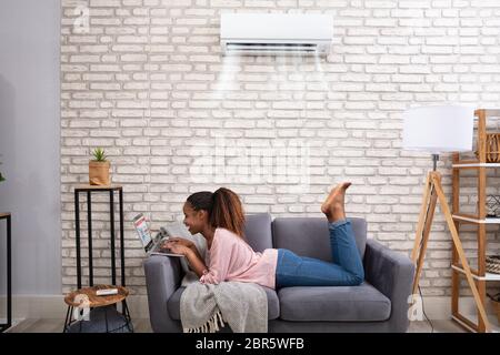 Happy Young African Woman Using Laptop Enjoying Cooling Under Air Conditioner At Home Stock Photo