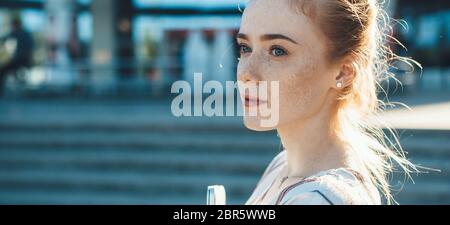 Freckled lady with red hair looking away while posing outside with a laptop in front of a sunset Stock Photo