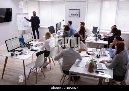 African Businessman Giving Presentation To His Colleagues Sitting On Table With Computers In Office Stock Photo